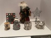 Various Christmas decorations including fabric Santa figurine, wooden Christmas tree frame, knitted snowman, glass candle holder with snowflake designs, and metal candle ring with leaf details.