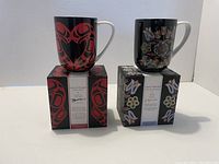 Two black coffee mugs with original decorative boxes featuring red heart design and colorful floral/butterfly patterns.