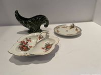 Three decorative dishes on a white surface: green cornucopia-shaped ceramic piece, leaf-shaped divided dish with floral pattern, round dish with handle.