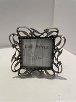 Close-up photo of a silver metal decorative photo frame with floral and swirl design holding a 3x3 inch placeholder photo with 'LIFE STYLE' text.