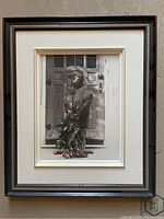 Framed black and white print showing a boy with flowers behind his back standing in front of a door, framed with dark wood frame and light matting.