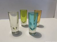 Four tall shot glasses in different colors: clear, lime green, blue, and amber. Thick clear bases visible.