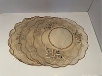 Four transparent oval place mats with scalloped edges and embroidered beige and brown flowers placed partially overlapping.