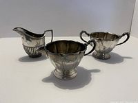 Photo of three silver-plated items including a creamer pitcher with ribbed base, a sugar bowl with scalloped rim and handles, and another vessel.