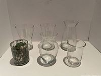Six vases displayed against a white background: five clear glass vases of varying sizes and one distressed silver vase with faux greenery.