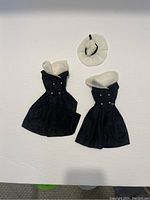 Two vintage Barbie black dresses with white collars and buttons, and one white hat with a black band, laid flat on white background.