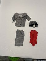 Top view of the four pieces of vintage Barbie clothing including a gray jacket, gray skirt, red bodysuit, and gray trimmed hat.