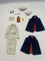 Photo displaying two navy-blue capes with red lining, white nursing dress, two nurse hats, black glasses, diploma, spoon, and brown and yellow bottles laid on a white background.