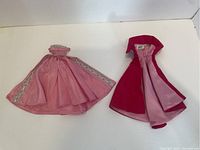 Two vintage Barbie clothing pieces laid flat on a white background showing the pink cape with silver trim and the red and pink dress/coat.