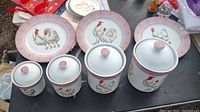 Four porcelain canisters with lids and rooster and hen illustrations, three plates with matching rooster and hen designs and red checked rims on a black table surface