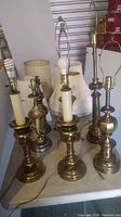 Seven brass lamps on table, mixed styles, varying heights, some with cream-colored faux candle sleeves and harps, with lampshades behind.