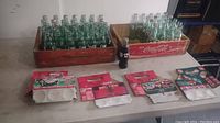Two wooden crates each holding multiple empty glass bottles (Coca-Cola and Sprite), four unopened flat cardboard display cartons, and one full Cal Ripken commemorative Coke bottle placed in front on table.