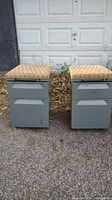Photo showing two heavy duty metal file cabinets side by side outdoors on pavement. Each cabinet has two drawers, gray metal finish, cushioned cloth tops with yellow-beige plaid pattern, and casters.