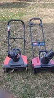 Front view of two small snow blowers with red bases and black chutes, sitting on grass.