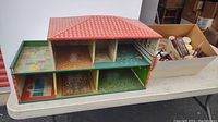 Photo of the vintage wooden doll house with multiple rooms, placed on white table alongside cardboard box of toy furniture.