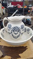 Full view of vintage ceramic pitcher placed inside matching wash basin showing blue floral garland pattern