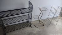 Three vintage metal plant stands/rack grouped together on a concrete floor including a black three-tier rack, a gold-tone decorative stand, and a white mesh shelf rack.
