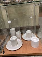 Photo of five Club Monaco hurricane/candle holders showing their white ceramic bases and clear glass cylindrical covers arranged on a wooden shelf.