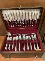 Photo of the silver plate flatware set arranged in open wooden box showing knives, spoons, forks, and ladle with red velvet interior.