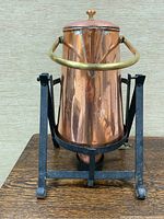 Front view of copper coffee server with brass handle on black metal stand