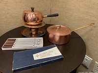 Copper fondue set with pot on burner stand and 6 fondue forks in box on table.