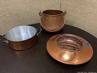 Photo showing three copper items on a dark surface: small copper pot with two handles, larger copper container with aluminum interior, and round copper flower arranger.