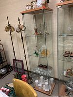 Tall glass display cabinet standing near a floor lamp, filled with various glassware and collectibles, showing full height and shelves.