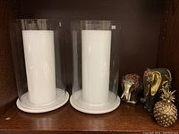 Two Club Monaco glass hurricane candle holders with large white pillar candles, two decorative elephants, and decorative pineapple trinket container arranged on a wooden shelf.