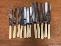 Set of 13 knives with cream-colored handles, metal blades showing maker marks, displayed on wooden surface.