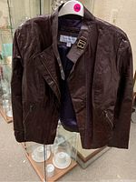 Front view of brown leather jacket hanging, showing zippered pockets, collar with buckle strap detail.