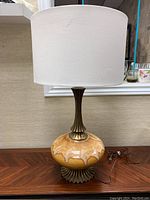 Full view of the lamp showing ceramic drip glaze base, copper-toned metal neck, and white fabric shade on wood table.