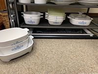 Multiple CorningWare dishes with blue cornflower pattern and some plain glassware pieces on shelf and floor