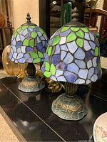 Pair of Tiffany style stained glass table lamps with purple, green, blue, and white glass shades on metal bases with textured greenish finish and power cords visible.