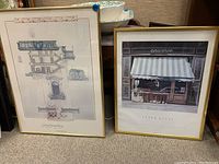 Both framed prints leaning against chair; includes architectural building print and storefront print with striped awning.