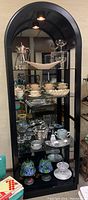 Front view of the black arched vintage display cabinet with glass shelves and accent light on top, containing various decorative items.