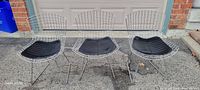 Three chrome wire chairs with black cushions side by side on driveway in front of garage.