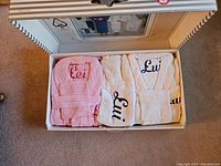 Photo of box containing folded bathrobes: one pink labeled 'Lei' and three white and blue labeled 'Lui.'
