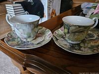 Set of two decorative espresso cups and matching saucers on a wooden surface, showing floral design with green leaves and white flower motifs, gold rim and handle accents.