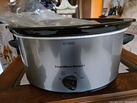 Front side view of Hamilton Beach slow cooker with control knob and lid, placed on wooden surface.