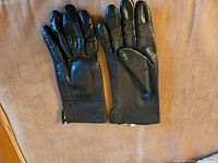 Pair of small women's black leather gloves laid flat on brown fabric background, showing both front and back sides with visible stitching and smooth finish.