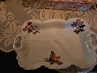 View of rectangular ceramic platter with two handles and multiple floral designs on a lace tablecloth.