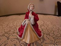 Porcelain figurine of a lady in a red cloak with fur trim and a yellow dress sitting with one hand on her chin.