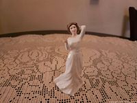 Porcelain figurine of a woman in a white dress posed with one arm raised, placed on a lace tablecloth.