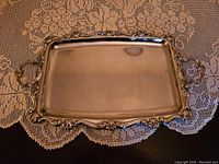 Rectangular silver plated serving tray placed on a lace tablecloth, showing decorative scrolling edges and handles, with visible surface tarnish.