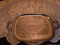 Rectangular silver plated serving tray with ornate handles and engraved pattern, placed on a lace tablecloth. Surface shows signs of tarnish and needs cleaning.