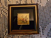 Photo of a small framed picture featuring a 23k gold plate with a nautical scene including a sailing ship and boats, mounted on a black matte background and wood-toned frame.