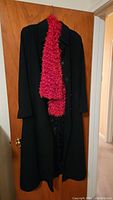 Long black wool coat hanging on a door, with a handmade red textured scarf draped around the collar area.