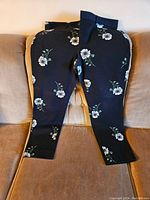 Black womens pants with white floral pattern laid flat on a beige cushioned surface.