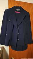 Navy blue women's pants suit blazer and pants hanging on wooden door, showing front of blazer with button details and pocket flaps.