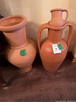 Photo showing two large terracotta vases and one smaller jug, all unglazed and reddish-brown in color, on a carpeted floor next to a wall.